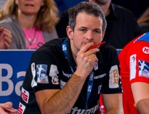 Ein Mann, der ein schwarzes Hemd mit Sponsorenlogos trägt, sitzt nachdenklich da und hält einen roten Stift an seinen Mund. Im Hintergrund sitzen Menschen, die leicht unscharf sind.