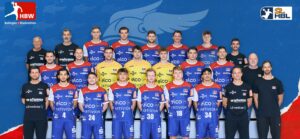 Eine Handballmannschaft der Männer posiert für ein Gruppenfoto in roten und blauen Trikots mit Sponsorenlogos, stehend und sitzend in Reihen vor einem blauen Hintergrund mit Team- und Ligalogos. Trainer und Betreuer sind in Schwarz gekleidet.