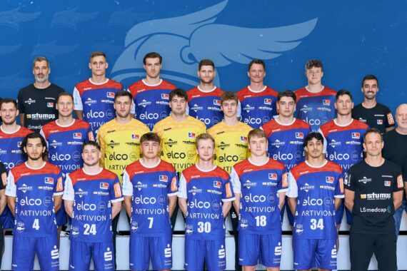 Eine Handballmannschaft der Männer posiert für ein Gruppenfoto in roten und blauen Trikots mit Sponsorenlogos, stehend und sitzend in Reihen vor einem blauen Hintergrund mit Team- und Ligalogos. Trainer und Betreuer sind in Schwarz gekleidet.