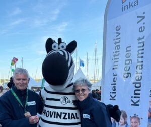 Zwei lächelnde Erwachsene stehen neben einem Zebra-Maskottchen bei einer Outdoor-Veranstaltung, dahinter ein Banner der Kieler Initiative gegen Kinderarmut e.V. und Boote - sie feiern die EHF-Europa-League-Saison 2025/2026 des THW Kiel Handball.