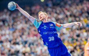 Ein männlicher Handballspieler in einem blauen Trikot mit der Nummer 30 springt und streckt seinen Arm aus, um den Ball während eines intensiven Spiels zu werfen, während im Hintergrund eine unscharfe Menschenmenge zu sehen ist.