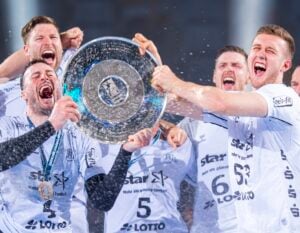 Eine Gruppe jubelnder männlicher Athleten in weißen Trikots hält jubelnd und lächelnd eine große silberne Trophäenplatte hoch, während Konfetti und Begeisterung in der Luft liegen.