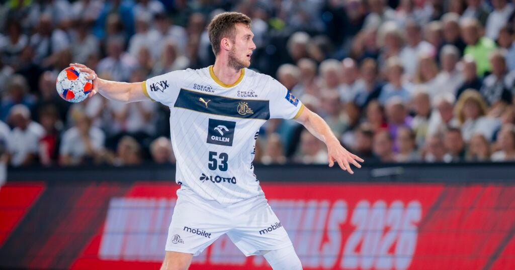HANDBALL 2025/26: THW Kiel - Rebud KPR Ostrovia Ostrow Wielkopolski Ein männlicher Handballspieler in einer weiß-marinefarbenen Uniform bereitet sich darauf vor, den Ball während eines Spiels zu werfen. Die Menge ist im Hintergrund verschwommen, und er hat seinen Arm erhoben, konzentriert auf die Aktion.