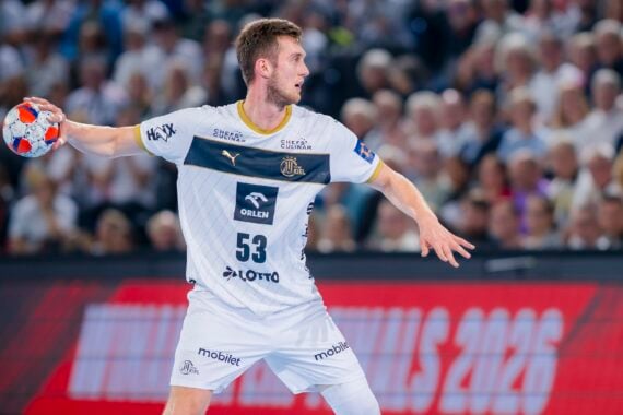 Ein männlicher Handballspieler in einer weiß-marinefarbenen Uniform bereitet sich darauf vor, den Ball während eines Spiels zu werfen. Die Menge ist im Hintergrund verschwommen, und er hat seinen Arm erhoben, konzentriert auf die Aktion.