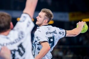 Ein Handballspieler in einem weißen Trikot bereitet sich darauf vor, den Ball während eines Spiels zu werfen, wobei der Arm eines anderen Spielers im Vordergrund zu sehen ist. Die Szene ist intensiv und actionreich.