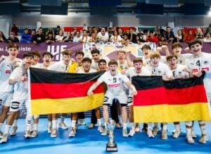 Eine Gruppe junger männlicher Sportler in weißen Uniformen feiert mit zwei großen deutschen Fahnen und einem Pokal, lächelnd und jubelnd vor einer Menschenmenge in einer Sporthalle.