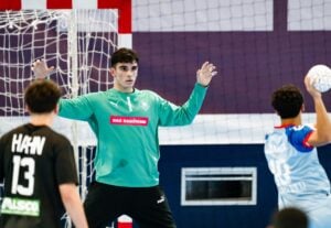 Ein männlicher Handballtorwart in einem grünen Trikot steht mit erhobenen Armen vor dem Tor, während sich ein gegnerischer Spieler zum Schuss vorbereitet. Ein anderer Spieler in einem schwarzen Trikot ist im Vordergrund zu sehen.