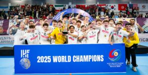 Ein jubelndes Jugendhandballteam in weißen Trikots feiert hinter einem großen blauen Schild mit der Aufschrift 2025 World Champions, mit jubelnden Fans und internationalen Flaggen im Hintergrund.