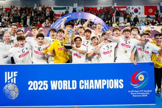 Ein jubelndes Jugendhandballteam in weißen Trikots feiert hinter einem großen blauen Schild mit der Aufschrift 2025 World Champions, mit jubelnden Fans und internationalen Flaggen im Hintergrund.