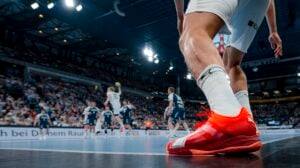 Eine Nahaufnahme des Beins und des leuchtend roten Schuhs eines Handballspielers auf einem Spielfeld, mit einer überfüllten Arena und anderen Spielern in Aktion im Hintergrund.