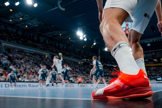 Eine Nahaufnahme des Beins und des leuchtend roten Schuhs eines Handballspielers auf einem Spielfeld, mit einer überfüllten Arena und anderen Spielern in Aktion im Hintergrund.