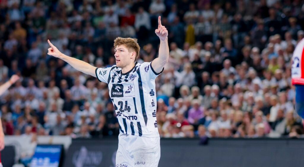 Ein männlicher Handballspieler in einem weiß-blau gestreiften Trikot mit der Nummer 24 hebt beide Arme und zeigt nach oben, während er auf dem Spielfeld mit einer verschwommenen, jubelnden Menge im Hintergrund feiert.