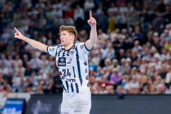 Ein männlicher Handballspieler in einem weiß-blau gestreiften Trikot mit der Nummer 24 hebt beide Arme und zeigt nach oben, während er auf dem Spielfeld mit einer verschwommenen, jubelnden Menge im Hintergrund feiert.