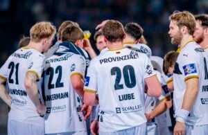 Eine Gruppe von männlichen Handballspielern in weißen Trikots mit schwarz-goldenen Verzierungen steht in einem Pulk zusammen, von hinten gesehen. Bei einigen sind Namen und Nummern zu sehen, und ein Spieler trinkt aus einer gelben Flasche.