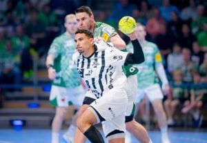 Ein Handballspieler in einem weiß-schwarzen Trikot bereitet sich darauf vor, den Ball zu werfen, während ein Spieler in einem grün-weißen Trikot ihn während eines Spiels eng verteidigt.