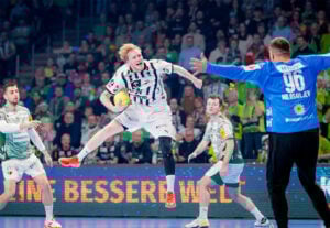 Ein Handballspieler in einem schwarz-weißen Trikot springt mit einem gelben Ball in der Hand vor einem blau gekleideten Torwart zum Schuss, während die gegnerische Mannschaft verteidigt und die Zuschauer von der Tribüne aus zusehen.