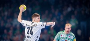Ein Handballspieler im weißen Trikot mit der Nummer 24 bereitet sich darauf vor, den Ball während eines Spiels zu werfen. Ein anderer Spieler in einem grünen Trikot schaut im Hintergrund inmitten einer unscharfen Menge zu.