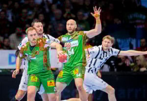 Vier männliche Handballspieler treten während eines Spiels gegeneinander an. Zwei Spieler in grünen Trikots halten den Ball und verteidigen, während zwei Spieler in weiß-schwarzen Trikots versuchen, den Ball zu blocken oder abzufangen. Die Zuschauer schauen aus dem Hintergrund zu.