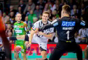 Ein Handballspieler in einem weißen Trikot wirft den Ball in Richtung Tor, während ein gegnerischer Torwart in einem schwarzen Trikot versucht, den Schuss zu blocken; im Hintergrund sind andere Spieler und Zuschauer zu sehen.