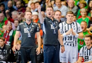 Ein Handballtrainer gestikuliert und ruft während eines Spiels Anweisungen, flankiert von Teammitgliedern und Spielern in schwarz-weißen Trikots, mit einer bunten, jubelnden Menge im Hintergrund.
