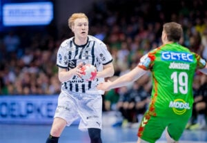 Ein Handballspieler in einem weiß-schwarzen Trikot hält den Ball, dicht bewacht von einem Gegner in einem grün-roten Trikot mit der Nummer 19. Das Spiel findet auf einem Hallenplatz statt, mit einer unscharfen Menschenmenge im Hintergrund.
