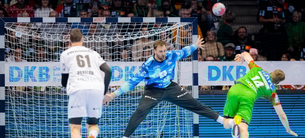 Ein Handballtorwart in Blau streckt sich, um einen Schuss eines Spielers in Grün abzuwehren, während ein anderer Spieler in Weiß im Vordergrund steht. Die Szene spielt sich in einer Halle mit einer jubelnden Menge ab.