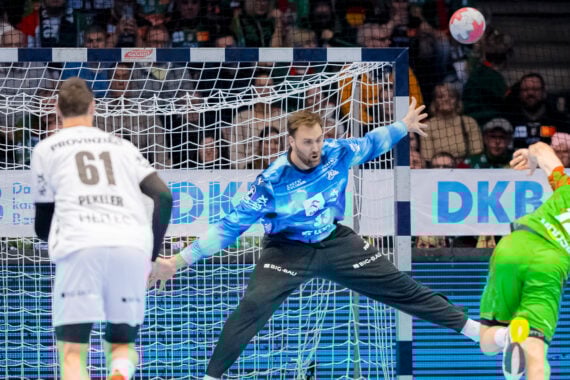 Ein Handballtorwart in Blau streckt sich, um einen Schuss eines Spielers in Grün abzuwehren, während ein anderer Spieler in Weiß im Vordergrund steht. Die Szene spielt sich in einer Halle mit einer jubelnden Menge ab.