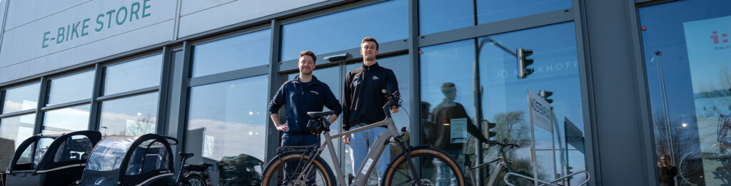 Zwei Personen stehen vor einem E-Bike-Geschäft neben einem modernen Elektrofahrrad, in dessen großen Glasfenstern Fahrräder und Zubehör ausgestellt sind.