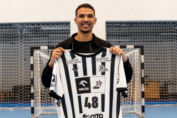 Ein lächelnder Mann steht drinnen auf einem Sportplatz und hält ein schwarz-weiß gestreiftes Trikot mit der Nummer 48 vor ein Tornetz.