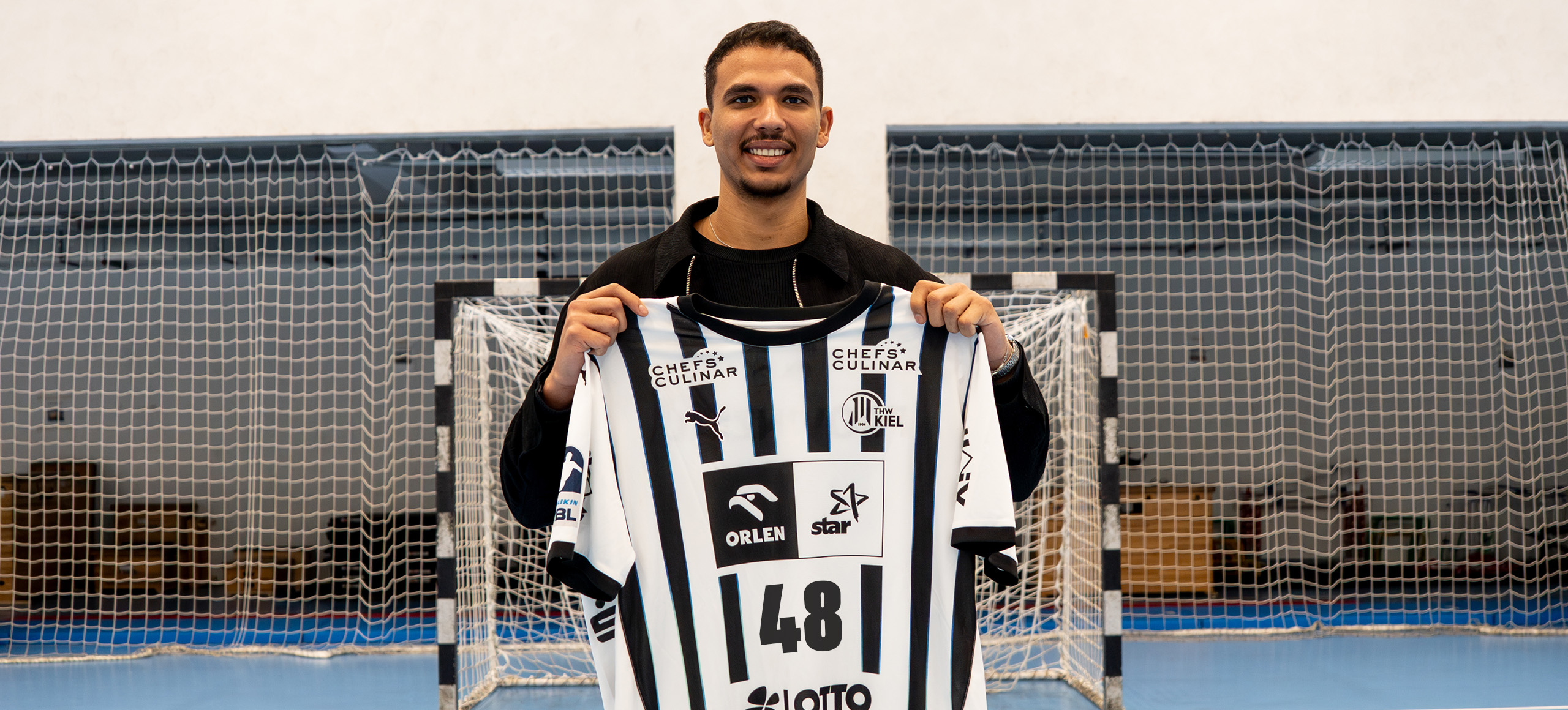 Ein lächelnder Mann steht drinnen auf einem Sportplatz und hält ein schwarz-weiß gestreiftes Trikot mit der Nummer 48 vor ein Tornetz.