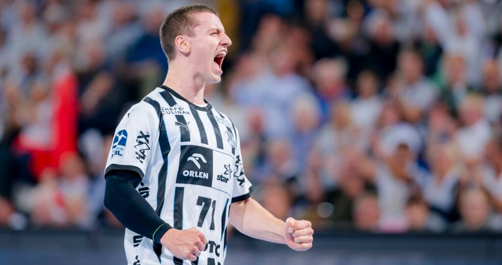 Ein männlicher Handballspieler in einem schwarz-weiß gestreiften Trikot schreit während eines Spiels aufgeregt mit geballten Fäusten. Die verschwommene Menschenmenge im Hintergrund suggeriert eine lebhafte Atmosphäre.