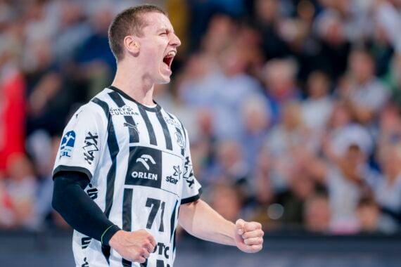Ein männlicher Handballspieler in einem schwarz-weiß gestreiften Trikot schreit während eines Spiels aufgeregt mit geballten Fäusten. Die verschwommene Menschenmenge im Hintergrund suggeriert eine lebhafte Atmosphäre.