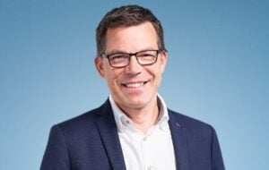 Ein Mann mit kurzen braunen Haaren und Brille, der eine dunkelblaue Anzugsjacke und ein weißes Hemd trägt, lächelt vor einem schlichten hellblauen Hintergrund.