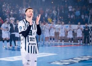 Ein Handballspieler in einem schwarz-weiß gestreiften Trikot steht auf einem blauen Spielfeld und klatscht mit seinen Mannschaftskameraden, die unscharf im Hintergrund zu sehen sind, was auf einen Moment der Anerkennung oder des Jubels während eines Spiels hindeutet.