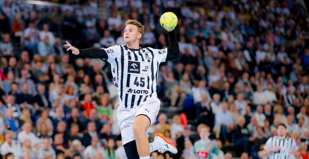 Ein männlicher Handballspieler in einem schwarz-weiß gestreiften Trikot springt in der Luft, hält einen gelben Ball und bereitet sich darauf vor, ihn zu werfen. Im Hintergrund ist eine unscharfe Zuschauermenge in einer Halle zu sehen.