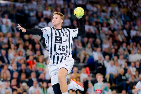 Ein männlicher Handballspieler in einem schwarz-weiß gestreiften Trikot springt in der Luft, hält einen gelben Ball und bereitet sich darauf vor, ihn zu werfen. Im Hintergrund ist eine unscharfe Zuschauermenge in einer Halle zu sehen.