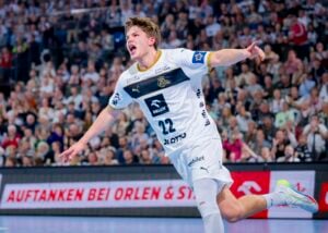 Ein männlicher Handballspieler in einem weißen Trikot mit der Nummer 22 jubelt mit ausgestreckten Armen auf dem Spielfeld, während eine große Menschenmenge auf der Tribüne im Hintergrund zuschaut.