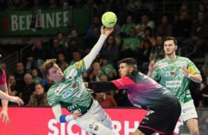 Zwei Handballspieler in Aktion: Der eine in einem grün-weißen Trikot streckt den Ball in die Höhe, während ein Verteidiger in einem schwarz-roten Trikot versucht, ihn zu blockieren. Im Hintergrund schauen Zuschauer zu.