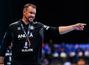 Ein männlicher Trainer in einem schwarzen Sportdress mit Sponsorenlogos zeigt während eines Spiels nach rechts und steht mit konzentrierter Miene am Spielfeldrand. Der Hintergrund ist unscharf, die Zuschauer sind sichtbar.