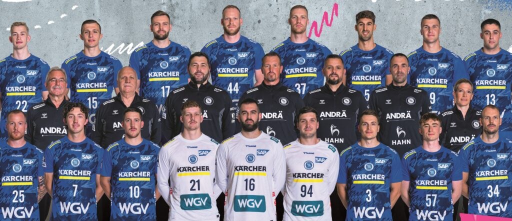 Eine Handballmannschaft der Männer posiert für ein Gruppenfoto in blau-weißen Trikots mit Sponsorenlogos. Die Spieler stehen in drei Reihen, Trainer und Betreuer in der mittleren Reihe, alle lächeln vor einem grauen und bunten abstrakten Hintergrund.