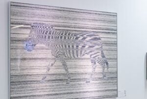 Ein gerahmtes Kunstwerk eines Zebras, das sich in einen Hintergrund aus horizontalen schwarzen und weißen Linien einfügt und so einen Tarnungseffekt erzeugt. Die Streifen des Zebras passen sich den Linien an, so dass sein Umriss teilweise verdeckt wird.