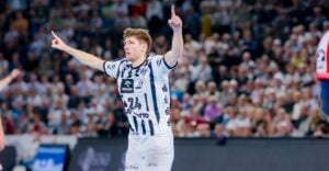 Ein männlicher Handballspieler in einem weiß-blau gestreiften Trikot mit der Nummer 24 hebt während eines Spiels beide Arme und zeigt nach oben, während im Hintergrund eine unscharfe Menschenmenge zu sehen ist.