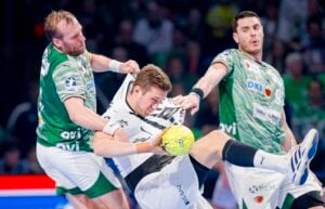 Drei männliche Handballspieler in Aktion; zwei in grünen Trikots verteidigen gegen einen in einem weißen Trikot, der einen gelben Handball hält. Die Szene ist intensiv und zeigt Körperkontakt und Anstrengung während des Spiels.