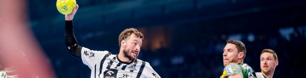 Ein Handballspieler in einem schwarz-weiß gestreiften Trikot springt und bereitet sich auf den Wurf vor, während zwei Spieler in grünen Trikots den Ball bei einem intensiven Hallenspiel verteidigen. Im Hintergrund sind Zuschauer und Schiedsrichter zu sehen.