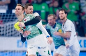 Zwei männliche Handballspieler in Aktion, wobei der eine in Grün den Ball festhält, während er von einem Spieler in Weiß eng verteidigt wird. Im Hintergrund sind zwei weitere Spieler und ein Tornetz zu sehen.