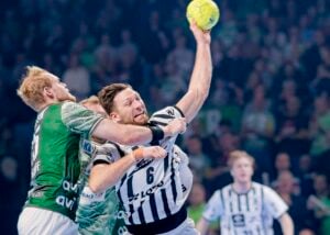 Ein Handballspieler in einem schwarz-weißen Trikot springt, um einen gelben Ball zu werfen, während er von einem Spieler in einem grünen Trikot während eines intensiven Spiels eng verteidigt wird, wobei die Zuschauer im Hintergrund verschwimmen.