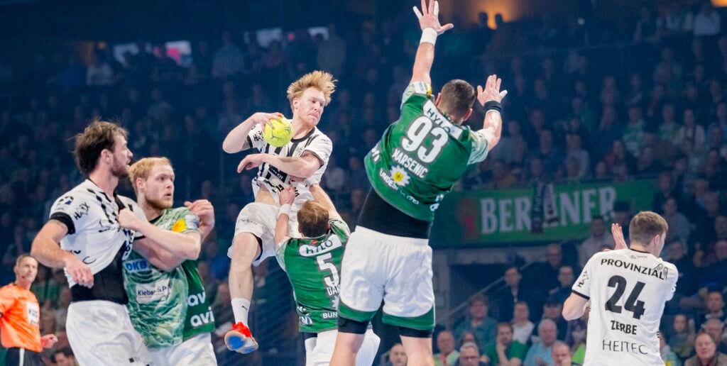 Ein Handballspieler in einem weißen Trikot springt in der Luft, um den Ball zu werfen, während er von gegnerischen Spielern in grünen Trikots umringt wird, die versuchen, ihn während eines intensiven Spiels zu blockieren.