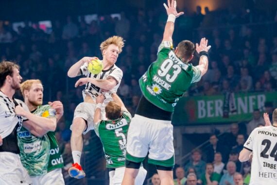 Ein Handballspieler in einem weißen Trikot springt in der Luft, um den Ball zu werfen, während er von gegnerischen Spielern in grünen Trikots umringt wird, die versuchen, ihn während eines intensiven Spiels zu blockieren.