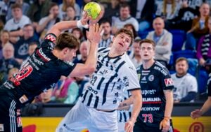 Zwei Männer in gestreiften und dunklen Trikots kämpfen während eines Spiels um einen Handball, wobei ein Spieler versucht, den anderen zu blockieren. Die Zuschauer schauen von den Tribünen im Hintergrund zu.