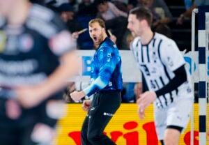 Ein männlicher Handballspieler in einem blauen Trikot schreit und ballt die Faust im Jubel, während Spieler in schwarzen und weißen Trikots in der Nähe auf einem Hallenplatz stehen. Im Hintergrund sind Zuschauer zu sehen.